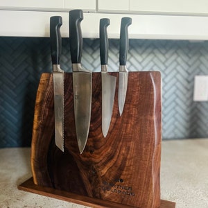May include: A wooden knife holder with a dark brown finish. The holder has a magnetic strip to hold knives and a base with the text "Partner Colorado" engraved on it. Five knives are attached to the holder.