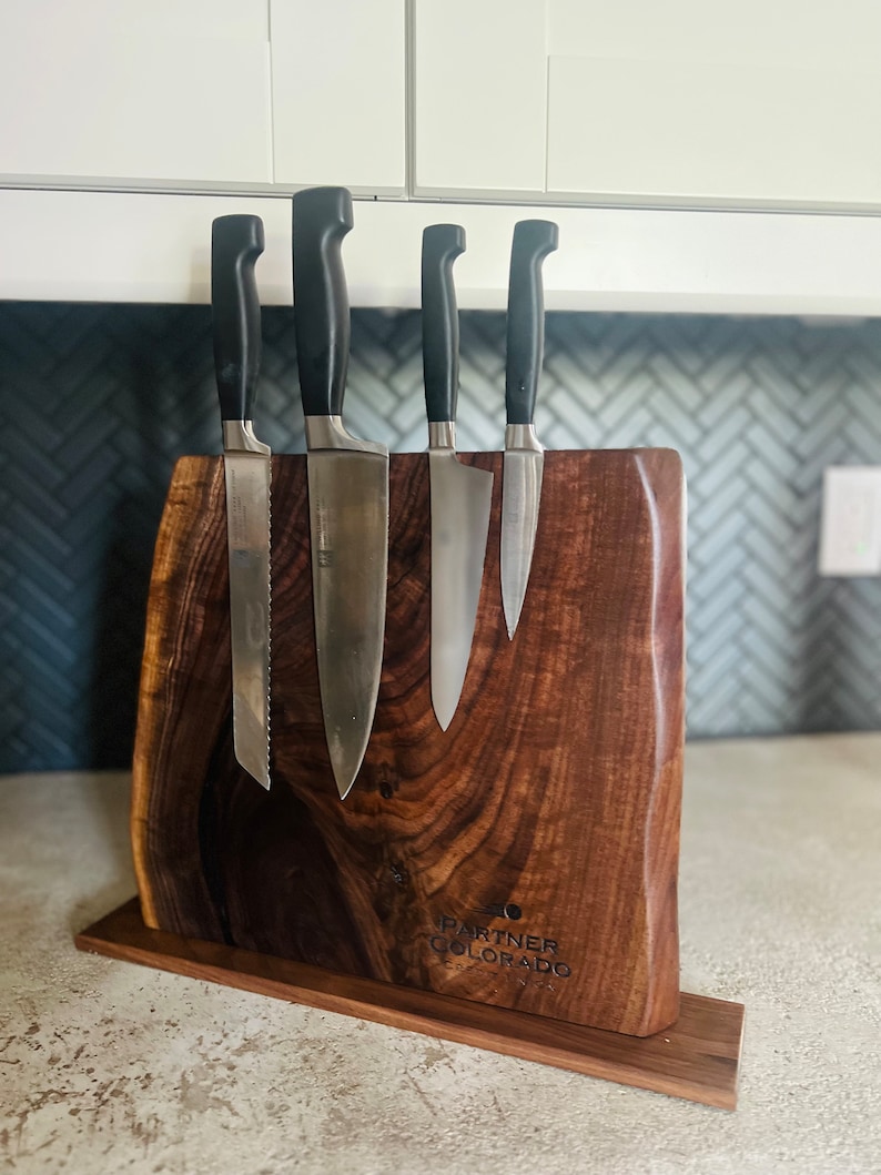 May include: A wooden knife holder with a dark brown finish. The holder has a magnetic strip to hold knives and a base with the text "Partner Colorado" engraved on it. Five knives are attached to the holder.