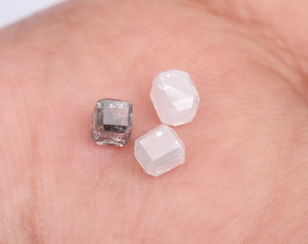 May include: Three rough-cut diamonds in varying shades of gray and white. The gemstones are held in a hand, showcasing their faceted surfaces and natural, unpolished appearance. The diamonds are small, with a cubic shape.