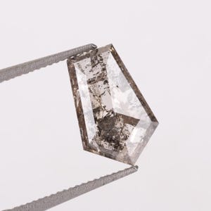 May include: A shield-shaped loose diamond featuring a salt and pepper appearance with prominent dark grey and black inclusions scattered throughout the translucent, faceted stone, held by a pair of metallic tweezers against a plain background.