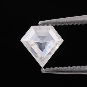 May include: A clear, square-cut gemstone held by silver tweezers against a black background. The gemstone has multiple facets, reflecting light and creating a brilliant appearance. The image highlights the clarity and shape of the gem.