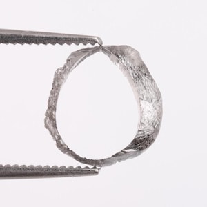 Pode incluir: Um anel de prata com superfície texturizada é segurado por uma pinça contra um fundo branco. O anel tem uma forma orgânica e irregular, sugerindo um design artesanal. O metal parece ligeiramente manchado, adicionando ao seu caráter único.