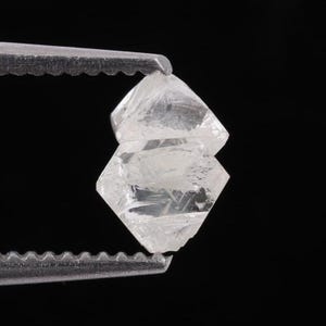 Op de afbeelding: Een ruwe, doorschijnende, kleurloze diamantkristal met een geometrische, octaëdrische vorm. De onbewerkte steen heeft scherpe randen, natuurlijke facetten en interne insluitingen, vastgehouden door een gekartelde metalen pincet.