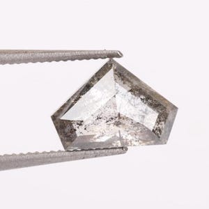 May include: A loose, shield-shaped salt and pepper diamond with a faceted surface. The gemstone displays a translucent, light gray body color with prominent dark gray and black inclusions scattered throughout the crystalline structure.