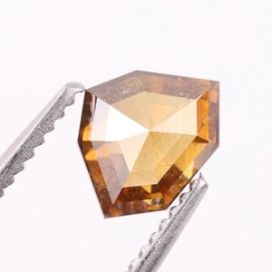May include: A loose, shield-shaped gemstone with a faceted surface and a warm, translucent amber-brown color. The stone is held by a pair of silver-toned metal tweezers with serrated edges, showcasing its geometric cut and reflective internal clarity.