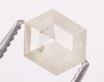 May include: A translucent, off-white, hexagonal-cut gemstone with visible internal inclusions and facets. The stone is held by two silver-toned metal tweezers, showcasing its geometric shape and pale, cloudy appearance against a plain background.
