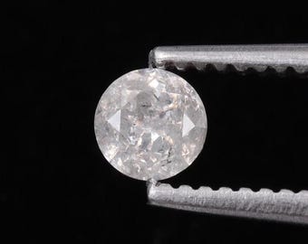 May include: A round, light-colored gemstone held by a pair of tweezers against a black background. The gem has a faceted surface with visible internal inclusions, giving it a unique, textured appearance. The gem is a neutral color, appearing white or light gray.