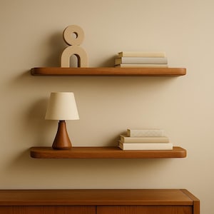 May include: Two wooden floating shelves with a warm brown finish are mounted on a beige wall. The top shelf holds a beige sculpture and a stack of books. The bottom shelf features a wooden lamp and a stack of books.