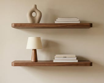 May include: Two floating wooden shelves with a neutral wall background. The top shelf holds a beige vase and a stack of books. The bottom shelf features a small lamp with a beige shade and a stack of books. The shelves are made of dark wood.