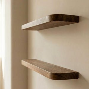 May include: Two floating wooden shelves with rounded corners are mounted on a neutral-colored wall. The shelves are a warm brown color, and the wood grain is visible. The shelves are evenly spaced apart, creating a minimalist design.