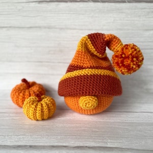 Puede incluir: Un gnomo de crochet con un gorro marrón, naranja y amarillo y un pompón. El gnomo está sentado sobre una superficie de madera blanca con dos pequeñas calabazas de crochet.