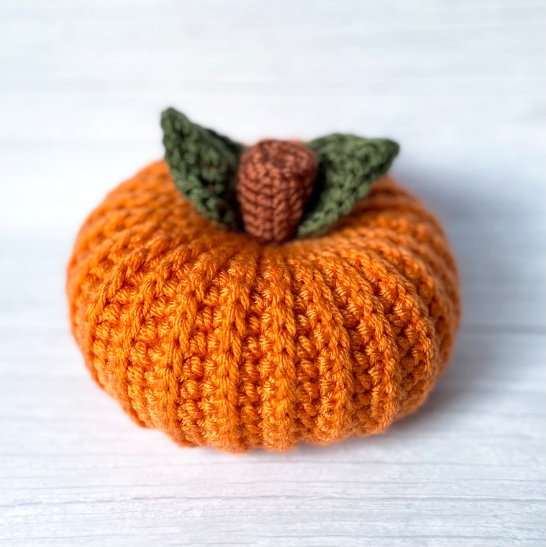 Op de afbeelding: Een gehaakte oranje pompoen met groene bladeren. De pompoen is gemaakt met een textuur-steek en heeft een bruine steel.