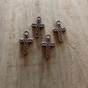 May include: Four small, copper-colored cross charms with a decorative openwork design. Each cross has a loop at the top for attaching to jewelry or other crafts. The crosses are arranged on a light brown wooden surface.