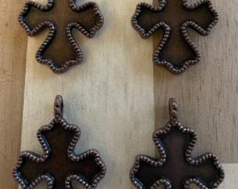 May include: Four bronze-colored cross pendants with a decorative beaded edge. Each cross has a loop at the top for attaching to a chain or cord. The crosses are displayed on a light brown wooden surface.