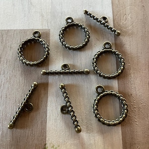 May include: Antique brass-toned toggle clasps and round ring clasps. The clasps have a textured, braided design. The toggle bars are straight and curved. The clasps are arranged on a light brown wooden surface.