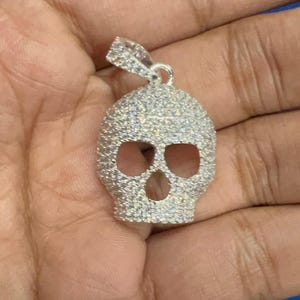 Puede incluir: Un colgante de plata con forma de calavera, incrustado con pequeñas piedras brillantes. La calavera presenta cuencas de los ojos y una cavidad nasal abiertas, con un lazo para una cadena. El colgante se sostiene en una mano.