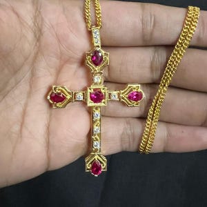 Könnte beinhalten: Ein goldfarbener Kreuzanhänger mit passender Kette. Das Kreuz hat rote Edelsteine an den Enden jedes Arms und in der Mitte, mit kleineren, klaren Steinen. Die Kette ist ein filigranes, gedrehtes Design.