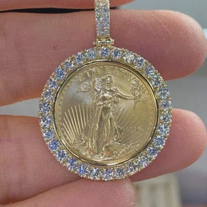 May include: A gold-toned circular pendant featuring a central coin with the embossed figure of Lady Liberty holding a torch and olive branch, surrounded by a border of clear, sparkling stones and a matching stone-encrusted bail at the top.