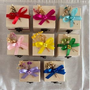 Puede incluir: Siete pequeñas cajas de madera con tapas con bisagras, cada una adornada con una colorida cinta de satén y flores secas. Las cintas son rojas, rosas, turquesas, amarillas, verdes, moradas y azules. Las cajas están dispuestas sobre una superficie blanca.