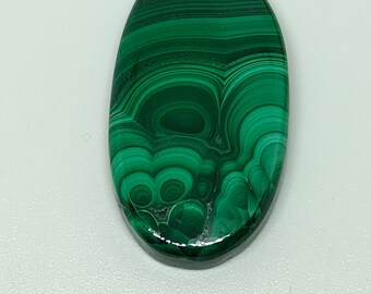 May include: Oval-shaped malachite gemstone with a polished surface. The gemstone features a vibrant green color with swirling patterns and bands of lighter and darker shades. The stone is set against a white background.