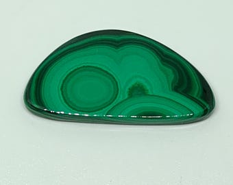 May include: A polished, oval-shaped malachite stone. The stone features concentric bands of dark and light green, creating a swirling pattern. The surface is smooth and reflective, with a glossy finish. The overall shape is reminiscent of a slice of a larger stone.