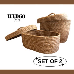 Peut inclure: Un ensemble de deux paniers de rangement ovales en corde tressée marron, chacun doté d'un couvercle assorti. Le texte WEDGO Living et SET OF 2 sont visibles sur l'image. Les paniers présentent un aspect tissé texturé de couleur beige.