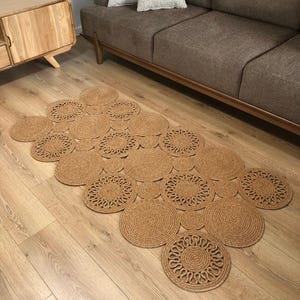 May include: A natural fiber rug composed of interconnected circles. The circles are arranged in a repeating pattern, with some featuring a decorative, openwork design. The rug is a warm, neutral brown color and rests on a light-colored wooden floor.