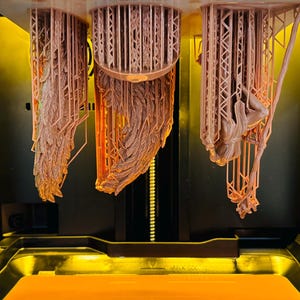 May include: Three light peach-coloured resin 3D printed figures hanging from a build plate inside a printer. The objects are suspended by a complex lattice of thin support structures, showing intricate details and textures created during the printing process.