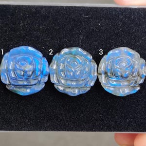 Puede incluir: Tres cabujones de rosas de piedras preciosas azules talladas. Cada cabujón es de un tono azul diferente con acentos grises. El diseño de la rosa es visible en cada piedra. Los cabujones se exhiben sobre una superficie negra.