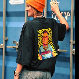 Könnte beinhalten: Schwarzes T-Shirt mit einer farbenfrohen Grafik einer Person mit Brille und Goldkette, mit dem Wort "RULEMAKER" auf dem Shirt. Die Grafik ist vor einem gelb-schwarzen Leopardenmuster-Hintergrund platziert. Die Person trägt eine orangefarbene Mütze.