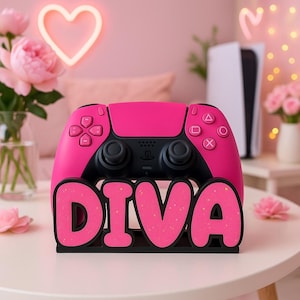 Könnte beinhalten: Ein pinkfarbener und schwarzer Videospiel-Controller liegt auf einem Ständer mit dem Wort "DIVA" in pinkfarbenen Buchstaben mit goldenem Glitzer. Der Controller befindet sich vor einem rosa Hintergrund mit einem Neonherz und Blumen.