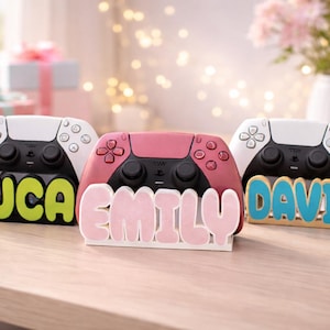Op de afbeelding: Drie gamecontroller-standaards, elk met een draadloze controller. De standaards zijn voorzien van gepersonaliseerde namen in dikke, verhoogde letters: LUCA in limoengroen, EMILY in zachtroze en DAVID in felblauw. De controllers zijn wit en zwart.