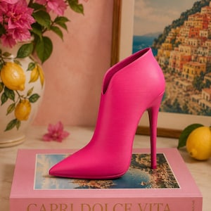 Könnte beinhalten: Eine leuchtend pinke Stiefelette mit Stiletto-Absatz, spitzer Zehenpartie und einem hohen, schlanken Absatz. Der Schuh hat eine glatte, strukturierte Oberfläche und einen tiefen V-förmigen Ausschnitt an der Vorderseite, was eine moderne Silhouette ergibt.