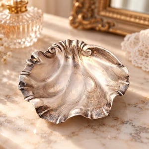 May include: A silver-toned metal dish shaped like a scalloped seashell with a polished, reflective surface and deep, curved ridges. The dish features a wide, undulating rim and a smooth, concave center, showcasing a metallic finish with subtle dark highlights.
