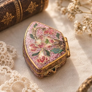 Pode incluir: Uma pequena caixa de metal de formato triangular com acabamento em esmalte rosa salpicado e um design floral em tons de rosa, verde e branco. A caixa possui uma estrutura de metal dourado, uma tampa articulada e um fecho ornamentado na frente.