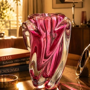 Könnte beinhalten: Eine dekorative Glasvase mit einem Wirbeldesign in Rosa- und Klartönen. Die Vase hat eine einzigartige, organische Form und steht auf einer Holzoberfläche. Der Hintergrund umfasst Bücher und eine Lampe, was auf eine Vintage-Ästhetik hindeutet.