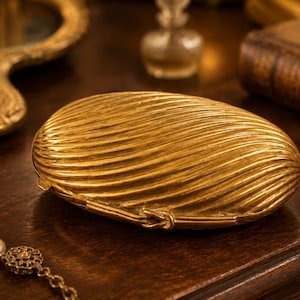May include: An oval-shaped gold-toned metal clutch with a textured, ribbed surface featuring diagonal lines. The accessory has a hinged opening mechanism with a small knot-shaped clasp detail on the front edge, creating a shell-like appearance.
