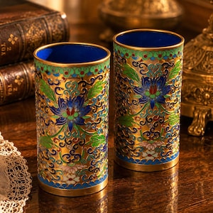 Pode incluir: Dois vasos cilíndricos com intrincados desenhos florais. Os vasos apresentam uma base dourada, um interior azul e são adornados com padrões florais azuis, verdes e vermelhos. O fundo inclui livros antigos e objetos decorativos.