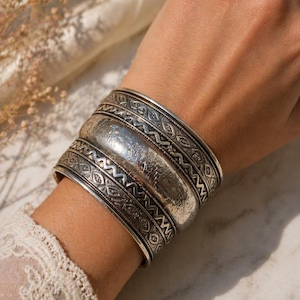 Könnte beinhalten: Ein breites silbernes Armreifarmband mit aufwendigen geometrischen Mustern und einem geprägten zentralen Design. Das Armband wird am Handgelenk getragen und zeigt seine detaillierte Handwerkskunst und seinen Vintage-Stil.