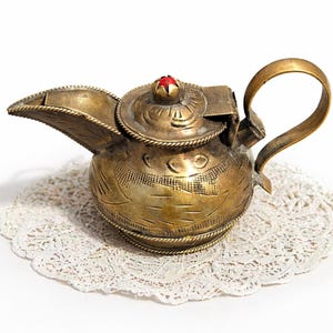 May include: A vintage brass teapot with intricate etched designs and a red gemstone on the lid. The teapot features a curved spout and a looped handle. It rests on a white lace doily, showcasing its ornate details and antique aesthetic.