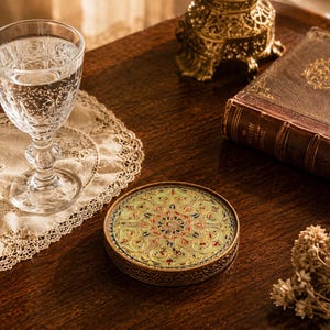 May include: A decorative coaster with a detailed floral design in red, gold, and blue sits on a dark wooden surface. A crystal glass filled with water and a vintage book are also visible.