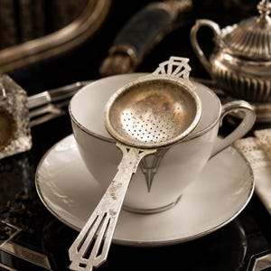 Könnte beinhalten: Ein Teesieb aus silberfarbenem Metall mit geometrischem Griffdesign liegt auf einer weißen Porzellantasse mit Untertasse. Die Tasse hat einen dünnen silbernen Rand und ein passendes geometrisches Muster an der Seite, ergänzt durch eine Zuckerdose.