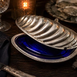 Puede incluir: Una fuente plateada en forma de concha con un interior azul oscuro. La fuente está abierta, revelando el cristal azul. Los bordes de la fuente tienen un diseño de cuentas. El fondo incluye una mesa de madera oscura, un vaso y una vela.