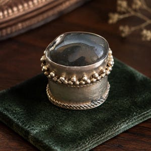 Op de afbeelding: Een zilveren ring met een grote, gladde, donkergrijze steen in een brede band. De band is versierd met een kralentextuur langs de bovenrand en een gedraaid touwpatroon langs de onderrand, wat zorgt voor een gedetailleerd en gestructureerd uiterlijk.