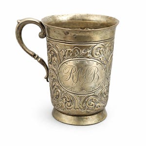 May include: Silver-colored mug with an ornate, engraved floral design. The mug features a handle and an oval cartouche with the engraved initials "RJB". The mug has a slightly flared rim and a small, circular base.