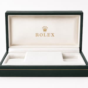 May include: An open, dark green Rolex watch box with gold trim. The interior is cream-coloured with a watch holder and the Rolex logo in gold. The box is empty against a white background.