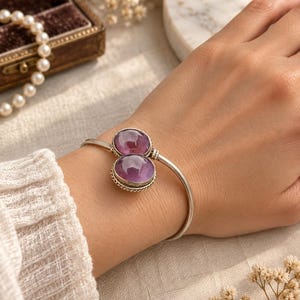 Op de afbeelding: Een zilveren armband met twee ovale, paarse amethist cabochon stenen gezet in een gedraaid metalen frame. De armband heeft een glad, open ontwerp dat om de pols rust en de gepolijste paarse edelstenen op subtiele wijze accentueert.