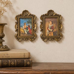 Può includere: Due cornici da parete rettangolari con bordi decorati color oro antico, caratterizzate da motivi a conchiglia nella parte superiore e inferiore. Ogni cornice contiene una piccola stampa dettagliata di una scena storica, una con due figure e l'altra con un bambino.
