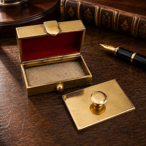May include: A rectangular brass box with a hinged lid and a small knob. The interior of the lid is lined with red fabric, while the base features a beige felt lining. A separate matching brass lid with a central knob sits beside the open box.