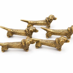 May include: Five brass dachshund figurines are displayed on a white background. The dogs have textured fur and collars. Each figurine is approximately 10 cm long, giving a vintage or antique appearance.